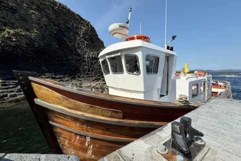 NTS A small boat with a wooden hull and a white cabin tied up at the new jetty. In the background, a cliff of hexagonal rock rises from the sea.