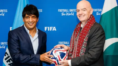A FIFA Syed Mohsin Gilani é uma camisa azul e à esquerda no blazer da Marinha. Infantino com um terno à direita e um xale vermelho no pescoço. Ambos estão segurando um futebol que está no meio deles. Possui linhas de ondas azul e vermelho. Há uma prancha escrita na palavra da FIFA por trás.