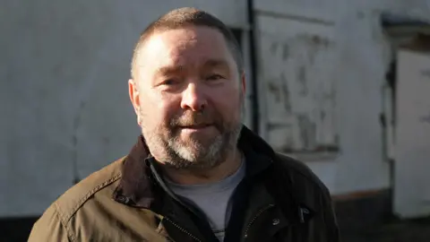 Shaun Whitmore/BBC Vic Freeman has cropped brown hair. He is wearing a green wax jacket and a grey T-shirt. He is standing outside the boarded-up building.