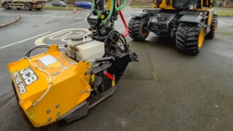 The image shows a large piece of road‑repair machinery on a street. The equipment is branded JCB in several places. In the foreground is an attachment with a bright yellow housing labelled JCB Pothole Pro, connected by hydraulic lines to a larger JCB vehicle behind it. The vehicle has four large tyres, an articulated arm, and a cab with clear windows. Traffic cones are placed along the road in the background, and several houses with driveways and hedges are visible. 