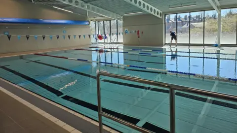 A large swimming pool with blue tiles. There is blue bunting hanging over the top of it. A person is standing next to the pool, mopping the tiles around it. The pool sits in front of a large set on windows which look out onto greenery and houses