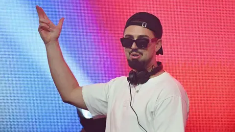 Getty Images Sonny Fodera is wearing a white T-shirt and black cap with black headphones around his neck. 