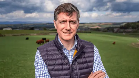 Simon Howie A man standing in a field, wearing a dark gilet. Various animals are grazing behind him. 