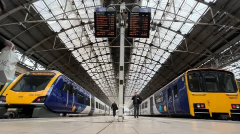 Network Rail Platform level image of the inside of Manchester Piccadilly railway station with two yellow, blue and white Northern trains. A couple of passengers are walking to and from the trains.