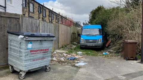 A metal bin on wheels next to a blue transit van parked in the alleyway.