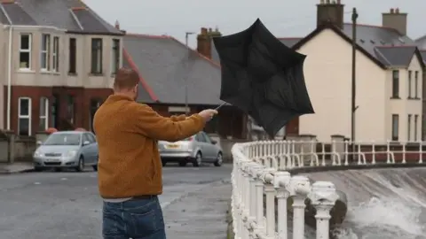 A man in a brown coat struggles with a black umbrella in Carnlough, County Antrim. There are white railings to his right and houses to his left.
