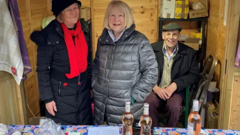 Marian McNeir Marian standing in a wooden chalet with another woman on the left and a man seated behind her. They are wearing warm winter clothing and smiling at the camera. The table in front of them has drink bottles and other goods laid out. 