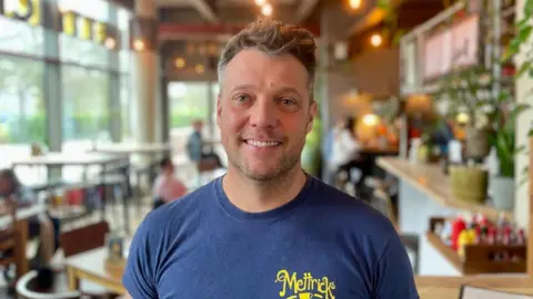 Spencer Bowman, a white man, standing in a coffee shop. He is wearing a blue t shirt with the shop's logo on it.