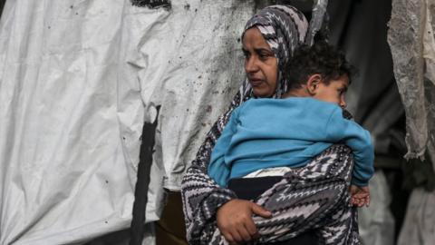 A Palestinian woman carries her child near makeshift tents after the Israeli shelling of a refugee tent encampment in al-Mawasi area west of Rafah, on 28 May 2024 in Rafah, Gaza