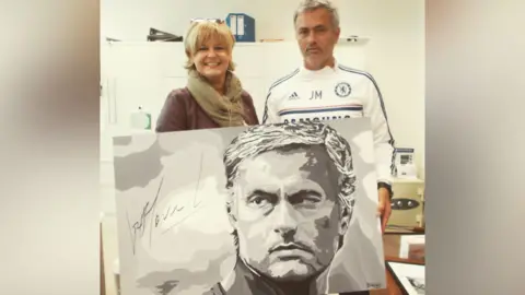 Sue Verity Art A woman in a beige scarf is standing next to Jose Mourinho, a man with greying hair, wearing a white sports top and holding a black and white portrait of himself