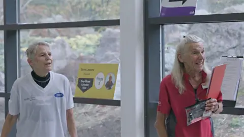 EVERYONE ACTIVE LEISURE The twins listening to someone speaking off camera to the right. Jennifer is laughing while holding a clipboard. She is wearing a red sports top, while her sister has a white top on.