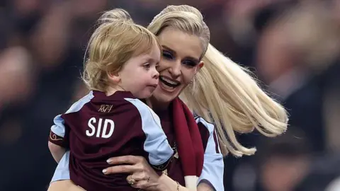 Getty Images Kelly Osbourne, who has long blonde hair that is waving behind her, is holding a young boy with blonde hair in her arms and smiling. The boy is wearing an Aston Villa shirt that says SID on the back