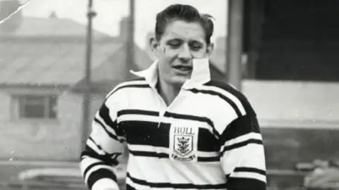 Hull FC A black and white photo of a man playing rugby league. He is wearing a black and white hooped shirt with a white collar. A badge on the chest depicts three crowns and has the word "Hull" in capital lettering.