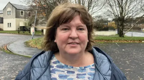 Niree McMorris has long brown hair and is wearing a white and blue top with a navy overcoat. She is standing in a car park, with a large white and grey building in the background.
