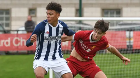 Getty An action shot on a football pitch showing Rogers, teenage boy with curly black afro hair in white shorts and a blue and white striped top is almost touching his opponent who is in a red kit. There is a goal behind them.