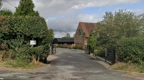 A driveway with two black gates, and hedges either side. Along the drive and to the right a red brick building can be seen, and further beyond that another building that is all clad in black metal