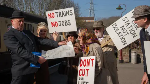 A group of protestors dressed in 1960s clothing carrying placards, harrassing a man dressed as Dr Beeching, who is looking at a railway map of the UK.