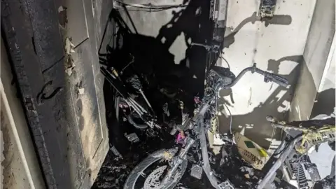 LFB The charred aftermath of an e-bike fire in a confined space. The frame of the bike is severely burned, with melted components and blackened walls around it. Debris and scorched materials are scattered on the floor.