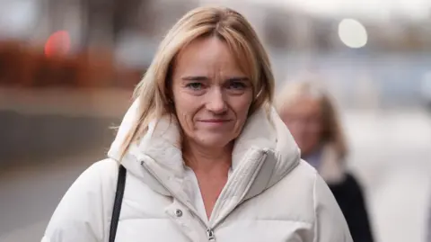 Sandie Peggie, a blonde woman wearing a white coat, arriving at court. She is staring straight at the camera and is expressionless.