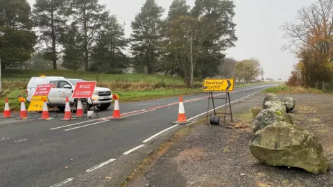 A white van is parked across the road behind a row of cones and a road closed and a diverted traffic sign. It's a rural location with lots of trees on either side of the road.