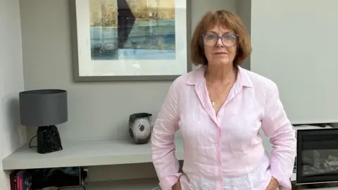 Cheltenham councillor Barbara Clark, wearing a pale top and standing at home in front of a painting on her wall.