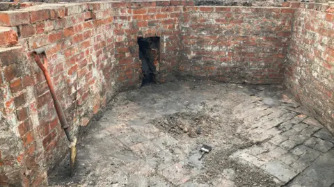 A dirty brick wall with a fireplace built into it. A shovel is leaning against the wall on the left of the image. The floor of the room is made up of old tiles but many of them in the centre are damaged