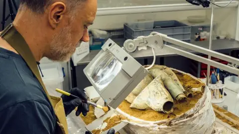 Norfolk Museums Service Jonathan Carr in a lab sitting at a table and looking through a magnifying glass on an arm as he brushed soil out of a bag holding a block of soil and Iron Age hoard items. 