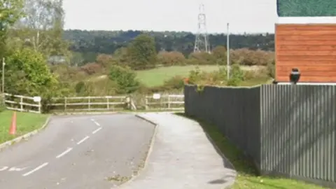General view of a road with a large expanse of land, trees and a electricity pylon on the background