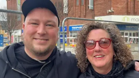 Andrew Turner/BBC Jordan Bishop, dressed in a black baseball cap, black teeshirt and black hoodie, stands in Great Yarmouth Market Place, with his left arm around his mother Tina's shoulder. She has long curly brunette hair, red rimmed glasses and is wearing a black scarf and puffa jacket.