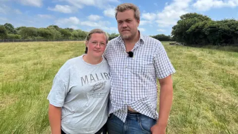 A man and woman stand side by side in an empty grassy field. Trees are visible in the background and there is a blue sky with clouds.