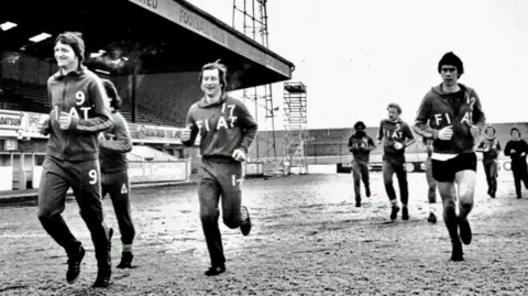 Cambridge United A black and white photo of nine Cambridge United players wearing tracksuits saying Fiat running at the Abbey Stadium in about the late 1970s or 1980s