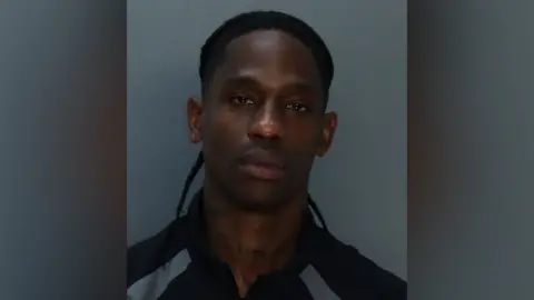 MDCR A mugshot of Travis Scott, his hair tied back. His expression is blank as he looks into the camera. He appears to be wearing a black and silver tracksuit top.
