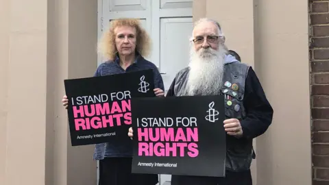 Jon Wright/BBC Jenny Brabazon and Mike Medhurst stand holding placards which read 'I stand for human rights'