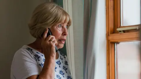 A woman with blond hair and a white patterned top holding a mobile phone to her ear and looking out of a wood-framed window