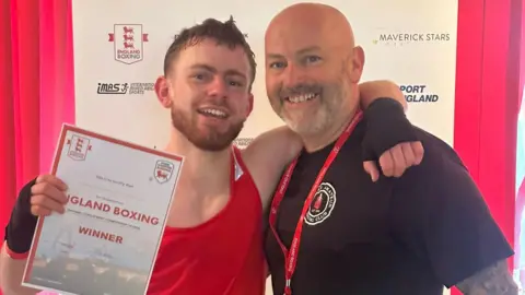 BBC Reuben smiling at the camera, wearing a red vest and black glove holding his winning certificate. His right arm is hung around is Dad Danny's neck, Danny is wearing a black t-shirt and smiling.