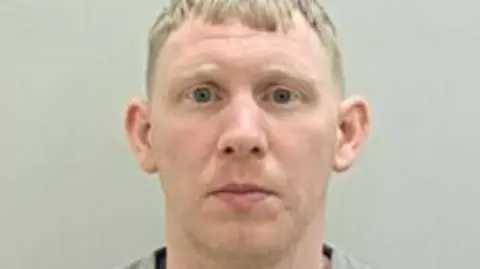 Police custody image of Ryan Gee who has blond hair and is wearing a grey top. 