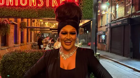 Roxy is standing outside on a street with bright neon signs behind her. She has her long brown hair up in a tall bouffant hairstyle with a turban style hair band. She is wearing full drag makeup and a glitzy necklace over a black fitted blazer.