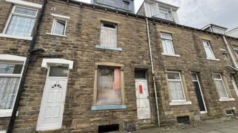 LDRS Row of stone-brick terraced houses on an overcast day. The central house appears abandoned or under renovation, with boarded-up windows and a worn white-painted door.