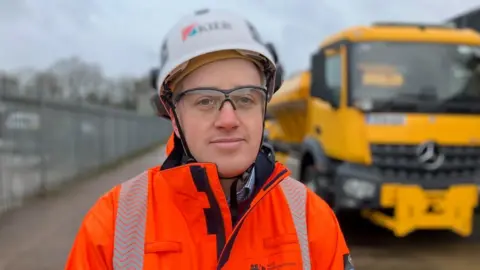 Alex Dunlop/BBC Allan Rigby, standing by an yellow gritting vehicle, looking at the camera, wearing a hardhat, safety goggles, an orange high-viz jacket, with a shirt underneath it. 