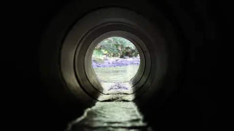 Getty Images View through a round concrete tunnel carrying a stream beneath a road - stock photo