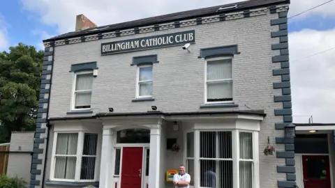 BBC / Ian Wood The exterior of the Billingham Catholic Club. The facade is mostly white with some blue accents. It has a bright red door in the middle and a brown door on the right hand side. The building is two storeys. Sean Rowbottom is standing, smiling at the foot of the building with his arms crossed.