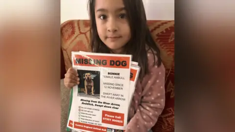 AJ Abrar A young girl with black hair wearing a pink jumper with white spots holds up a flyer titled "missing dog". 