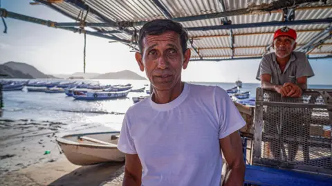 Fisherman Pablo Marín stands on the beach. He is wearing a white T-shirt and behind him, a man wearing a red baseball cap leans on a metal railing. Fishing boats can be seen on the water behind them.