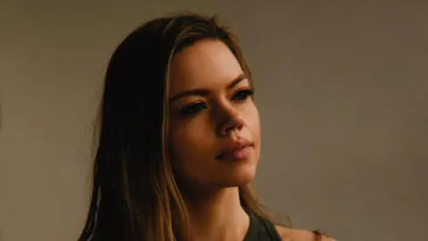 Eloise Skinner Eloise Skinner looks away from the camera. She has long dark blonde hair and wears a dark tank top. 