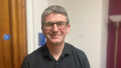 Photograph of Gavin White, the Executive Member for Housing at Manchester City Council. He is pictured at Manchester town hall. He has short grey hair, wears glasses, and is photographed in a dark grey open shirt.