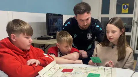 Emma Glasbey/BBC Two boys and a girl at Wincobank youth club look at a boardgame they've designed, with an adult also looking at the game