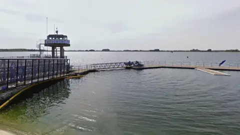 Google Farmoor Reservoir with a large jetty and lookout point pictured in the foreground. It is constructed of a metal framework and floating pontoons. The water is greenish blue. Trees on the other side of the reservoir are visible in the distance.