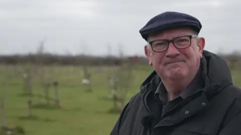 Bernard Pendleton wearing a blue flat cap, black rimmed glasses and black waterproof coat with the woodland behind him