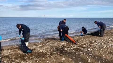 Sea Life Five people dressed in Navy blue uniforms are helping to release the seals back into the wild. They are standing on a beach by the water's edge with the seals in long carry nets.