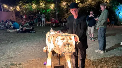A man in a black suit and hat is standing next to a dog made of paper and lit up from within. Festival-goers can be seen lounging on the floor in the background, with a few stopping to look at the man and his dog lantern.